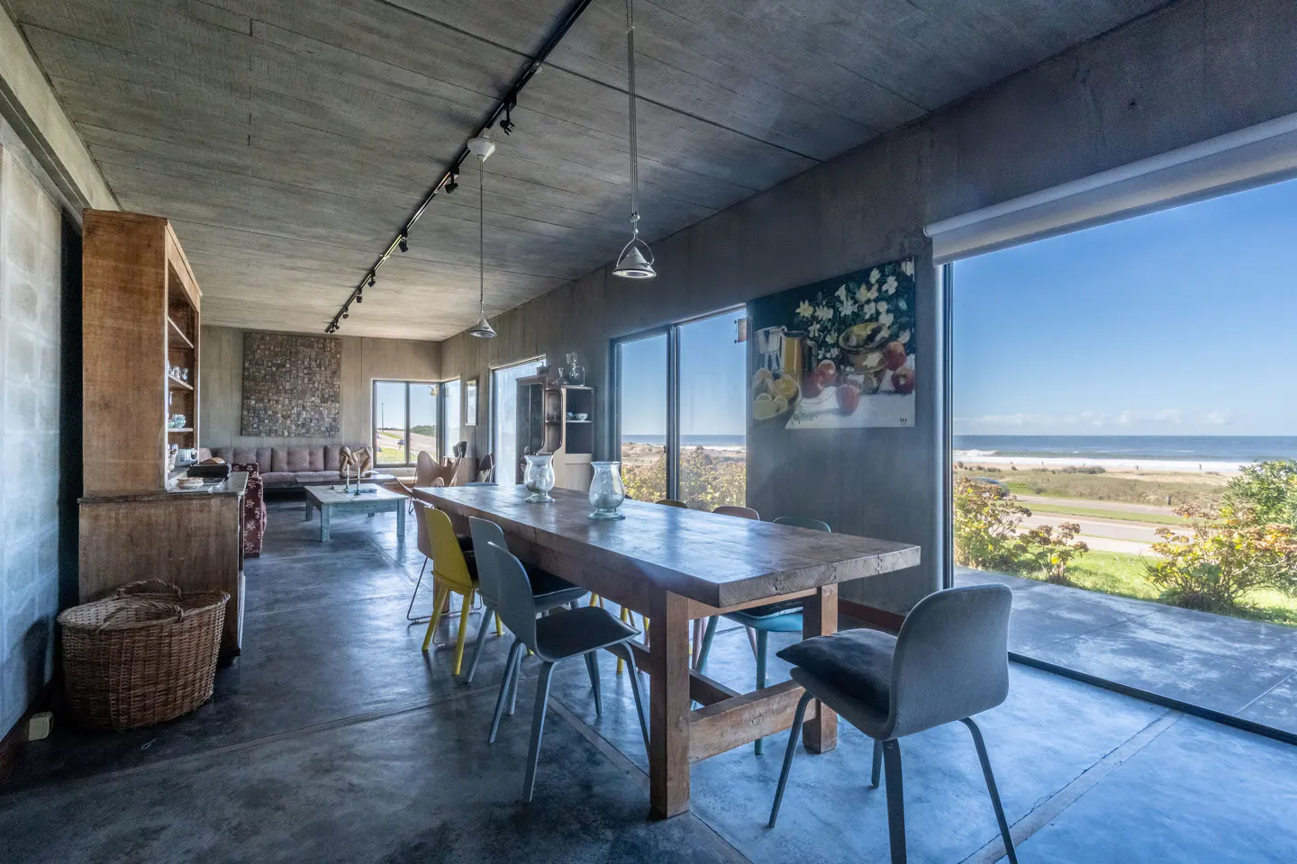 A modern dining room with a long wooden table and chairs, concrete walls, and a large window overlooking the ocean.