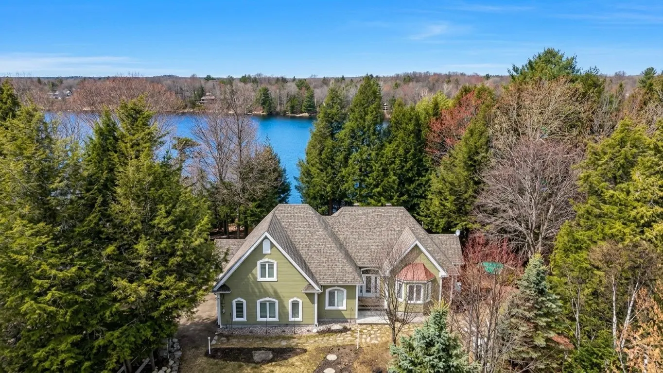 Aerial view of a green house with a gray roof, surrounded by trees, with a lake in the background.