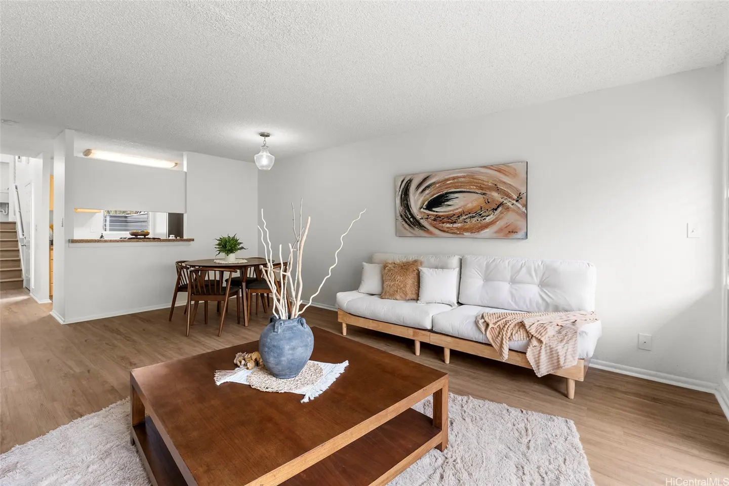 Bright living room with a white sofa, wooden coffee table, and a dining area with a table and chairs on wood-look flooring.