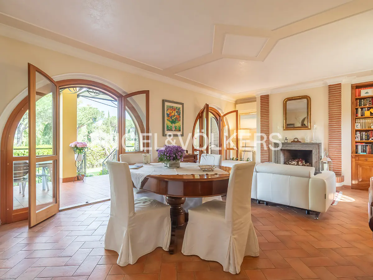 Bright living room with terracotta floors, a round table with white chairs, and arched doors leading to a balcony.