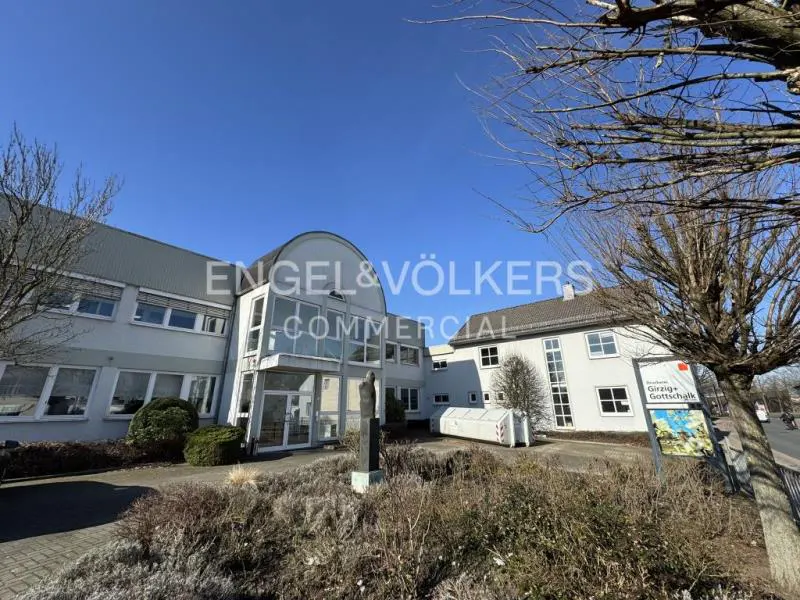 Exterior of a light gray commercial building with Engel & Völkers signage under a blue sky. A statue stands in front.