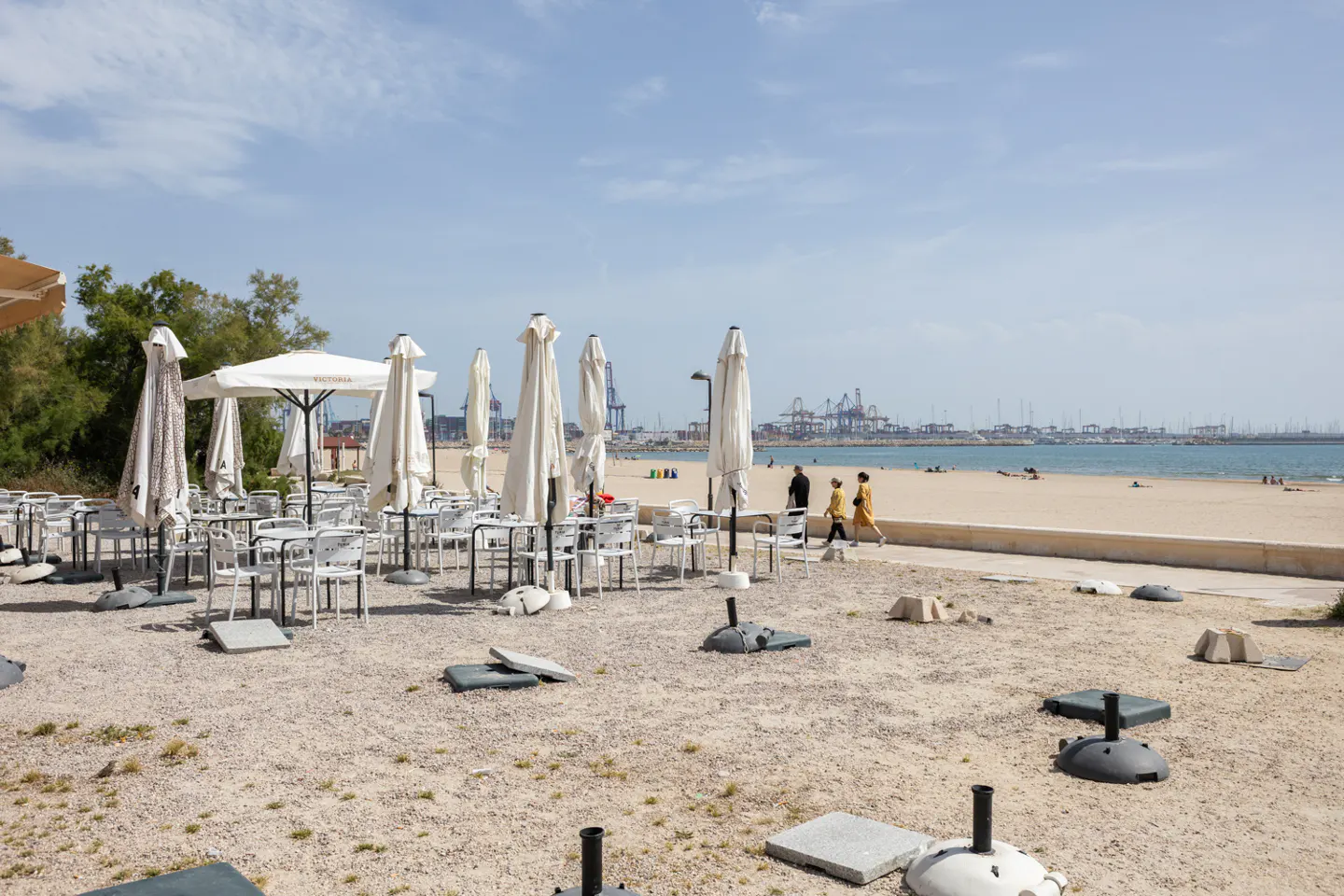 Beachside cafe with closed white umbrellas and empty white chairs. People walk on the beach in the background. Blue sky.