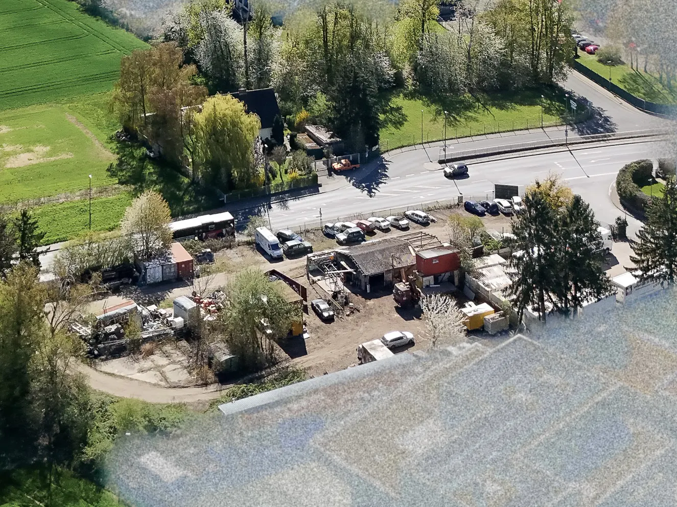 Aerial view of a property with a damaged building, parked cars, and construction equipment, near a road and green fields.