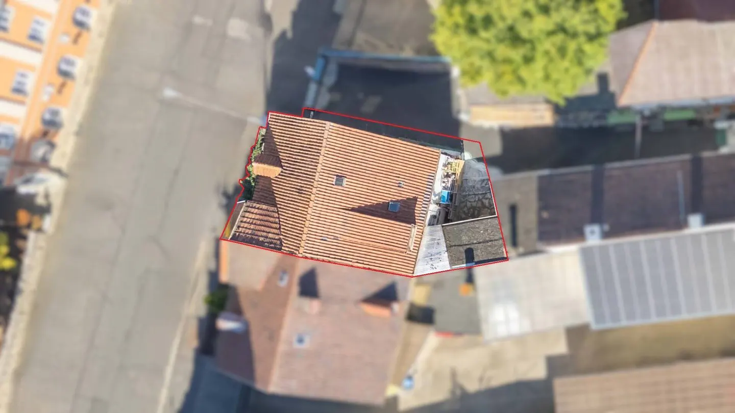 Aerial view of a building with a red tile roof, outlined in red, surrounded by other buildings and a street.