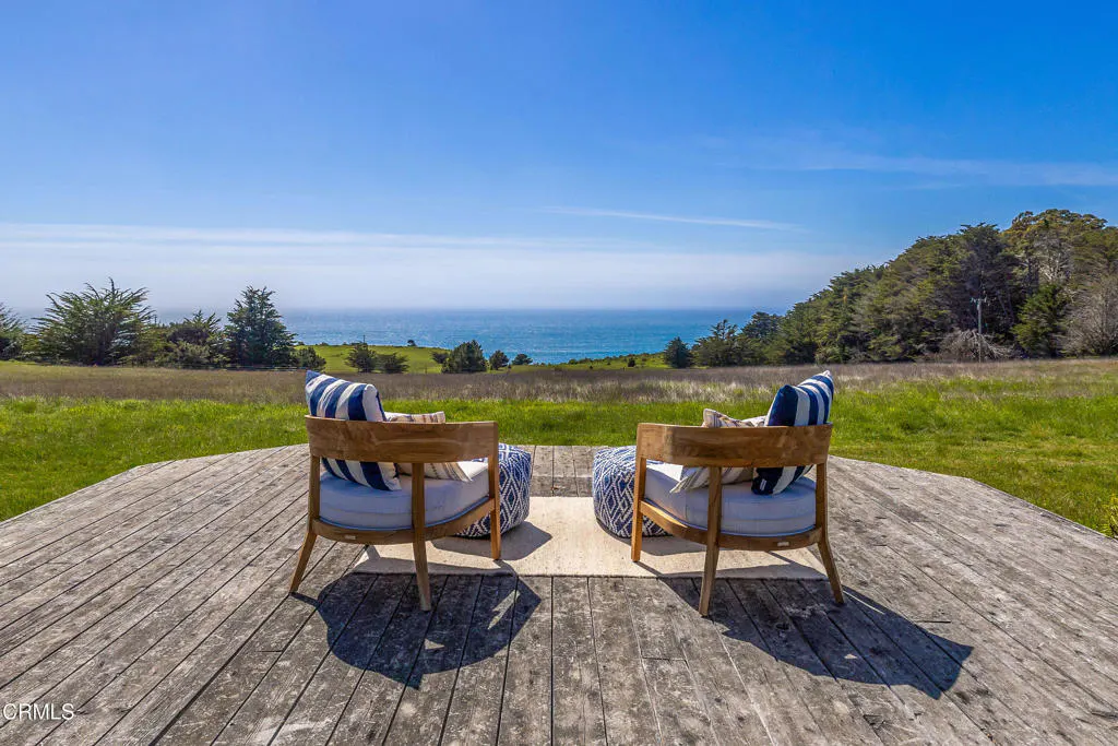 Two chairs with blue striped pillows face the ocean on a wooden deck overlooking a green field.