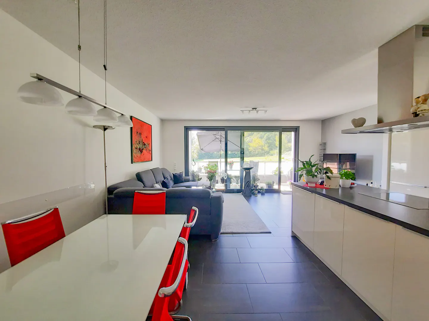 Open-concept living space with a white dining table, red chairs, a gray sofa, and a modern kitchen with white cabinets. Sliding glass doors lead to a balcony.