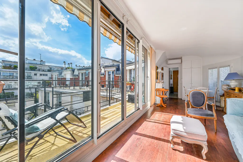 Bright living room with hardwood floors, blue chairs, and a balcony with lounge chairs overlooking city buildings.