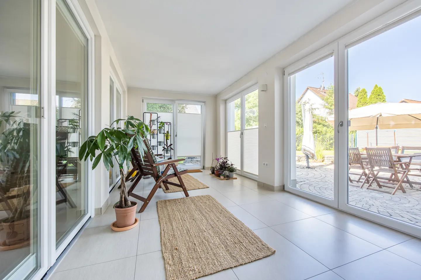 Bright sunroom with white tile floor, large windows, and two brown wooden chairs on jute rugs. A potted plant sits near the window.