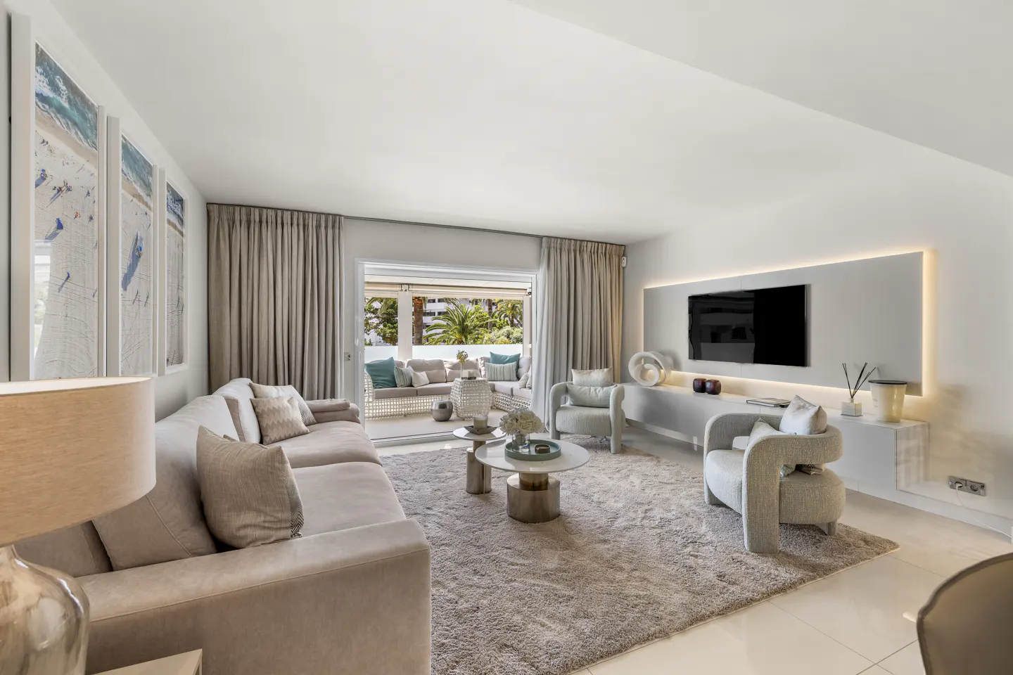 Bright living room with beige sofa, chairs, and rug. A TV is mounted on the wall with a light panel. Balcony with outdoor seating is visible.