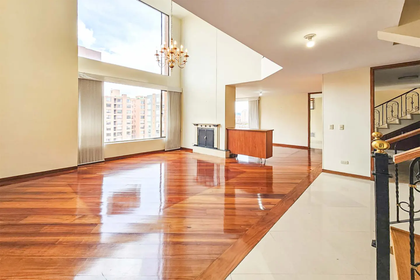 Bright, open living room with hardwood floors, a fireplace, and large windows overlooking a city. A chandelier hangs from the high ceiling.