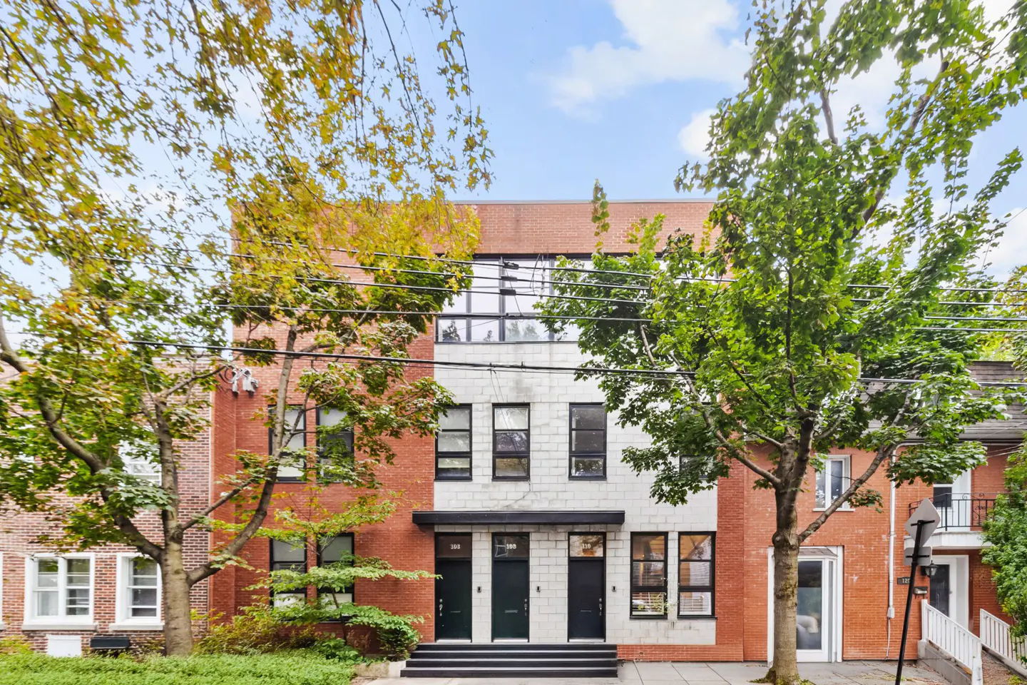 Exterior view of brick townhouses with black doors and windows, framed by green trees under a blue sky.