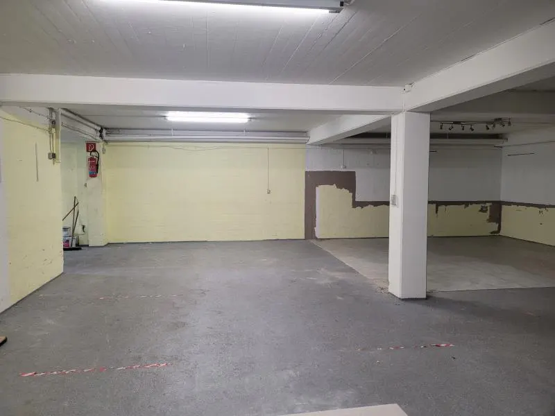 An empty, unfinished basement space with concrete floors, white support pillars, and pale yellow walls under bright lights.