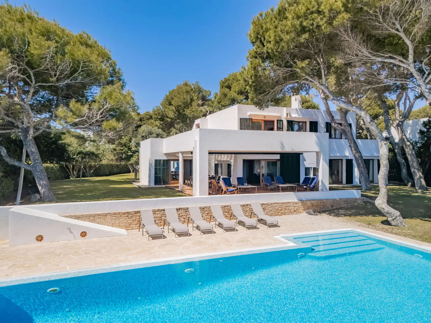 Modern white villa with a blue pool, lounge chairs, and green trees under a clear blue sky.