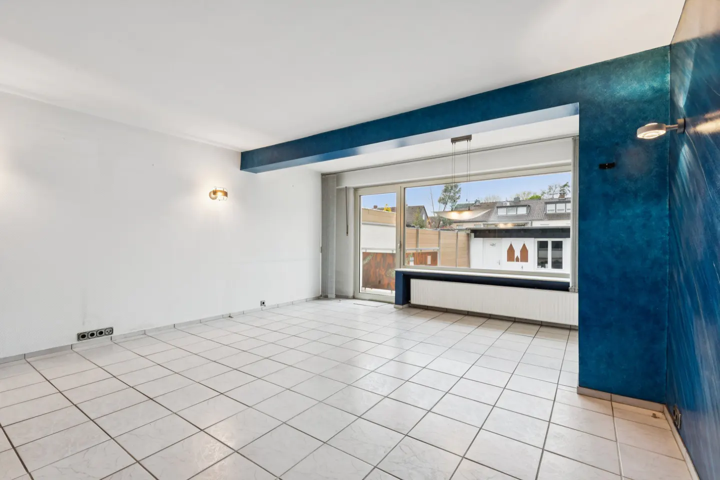 Bright, empty room with white tile floor, white walls, and a large window. One wall is blue with a modern light fixture.