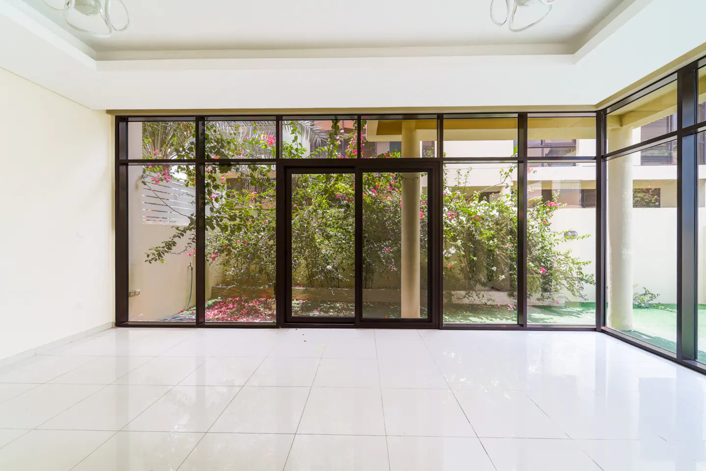 Bright, empty room with white tile floor and large windows. Outside, lush greenery and pink flowers are visible.