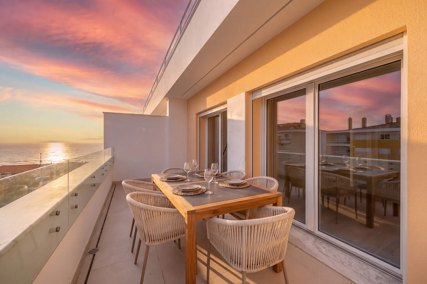 Balcony with a dining table set for six, overlooking the ocean at sunset. Wicker chairs surround a wooden table with plates and glasses.