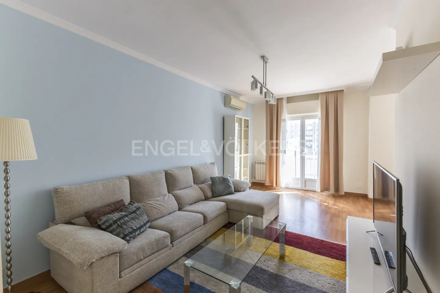 Living room with a beige sectional sofa, glass coffee table, and a colorful striped rug. Light blue walls and hardwood floors.