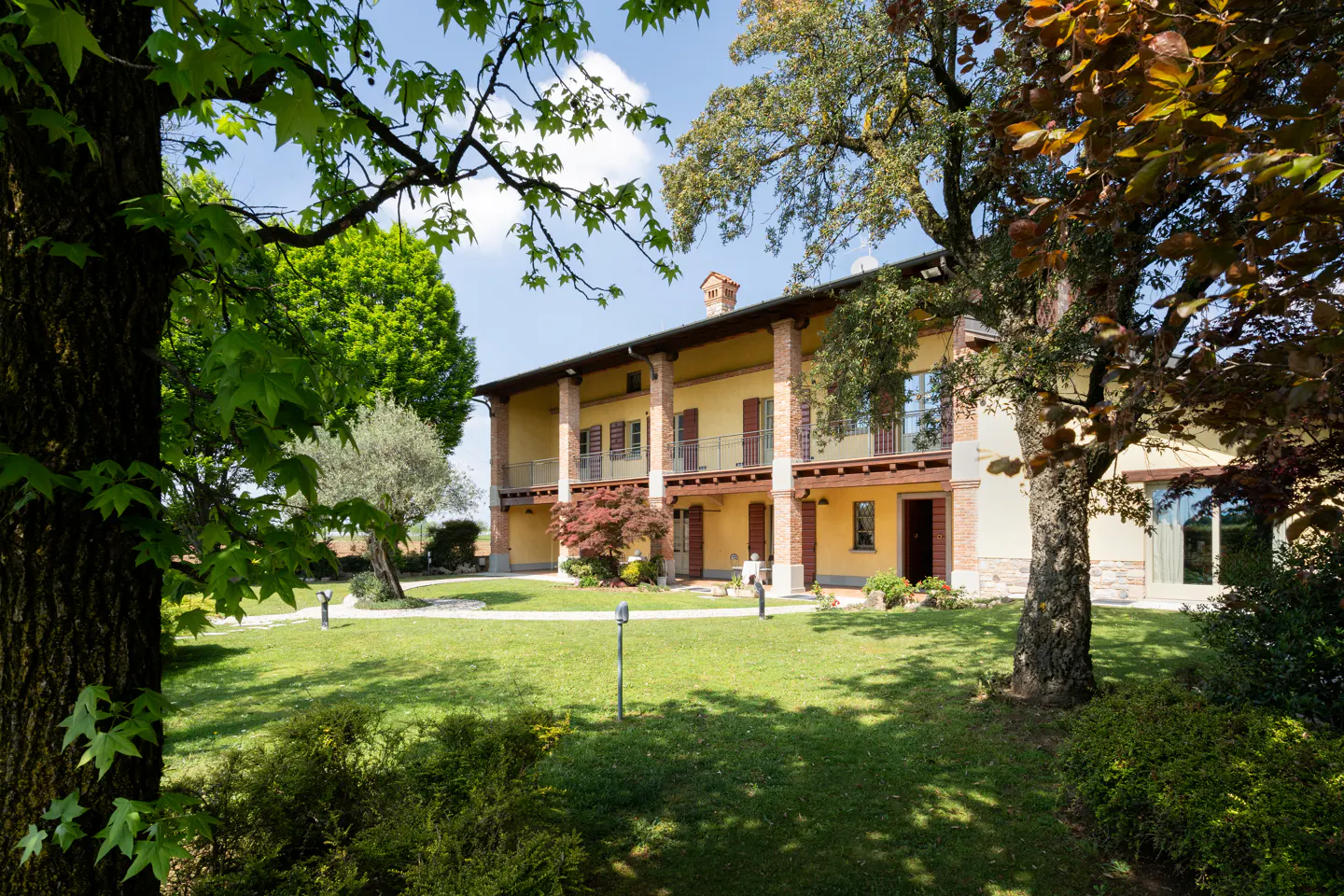 Two-story yellow house with brick columns and brown shutters, surrounded by green lawn and trees under a blue sky.