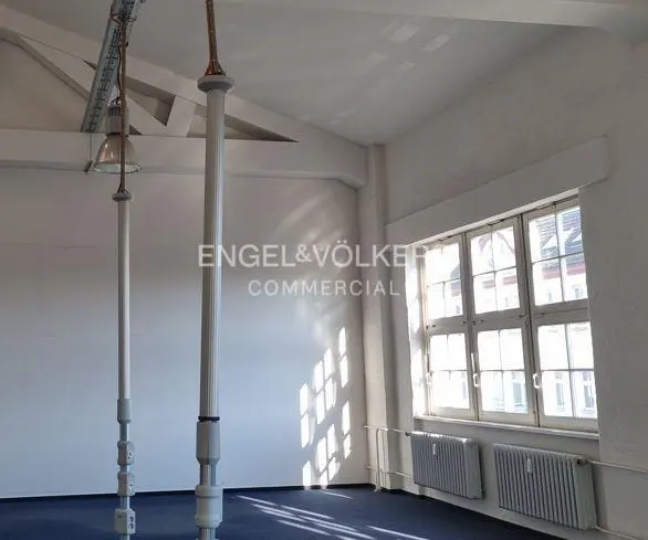 Bright, empty commercial space with white walls, blue carpet, tall white support columns, and large windows with radiators below.