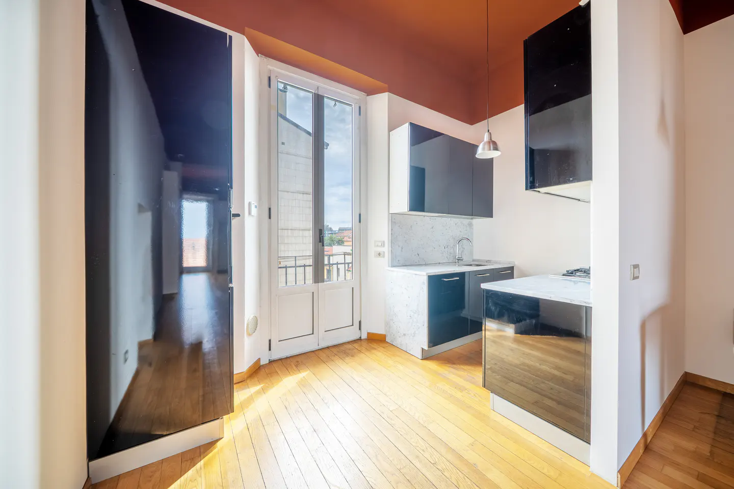 A modern kitchen with black cabinets, white countertops, and a marble backsplash. A white door leads to a balcony. The ceiling is painted brown.