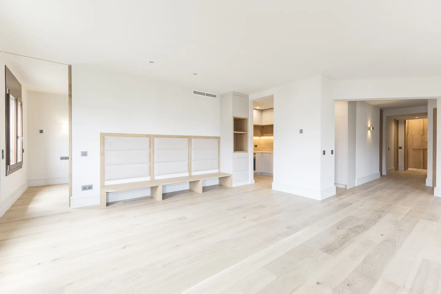 Bright, minimalist room with light wood floors, a built-in bench seat, and open doorways leading to a kitchen and hallway.
