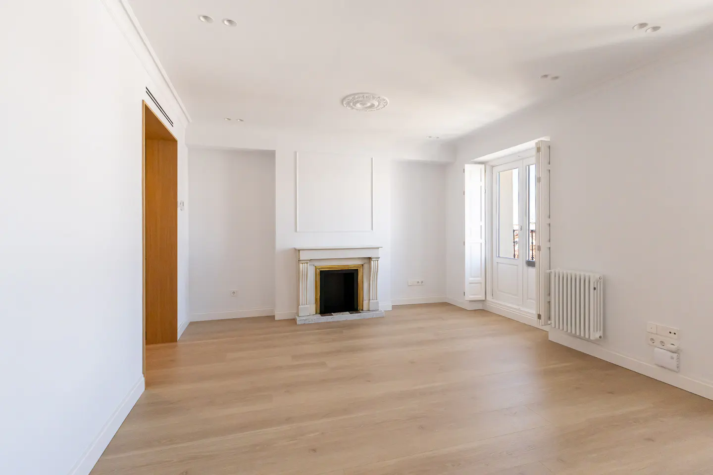 Bright, empty room with light wood floors, white walls, and a marble fireplace. A white door leads to a balcony.