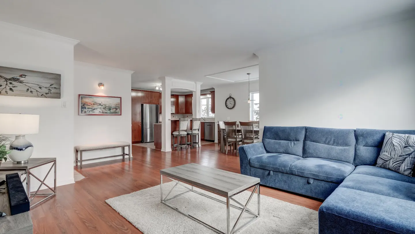 Open-concept living space with a blue sectional sofa, wood floors, and white walls. Kitchen and dining area visible in the background.