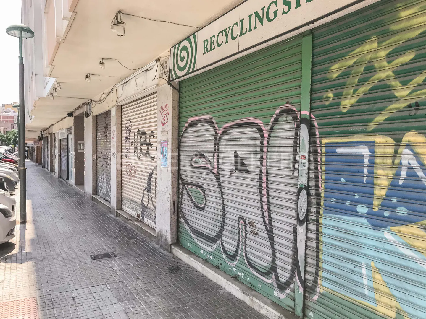 A street view of closed storefronts with graffiti on metal shutters. "Recycling" sign visible. Cars parked along the street.