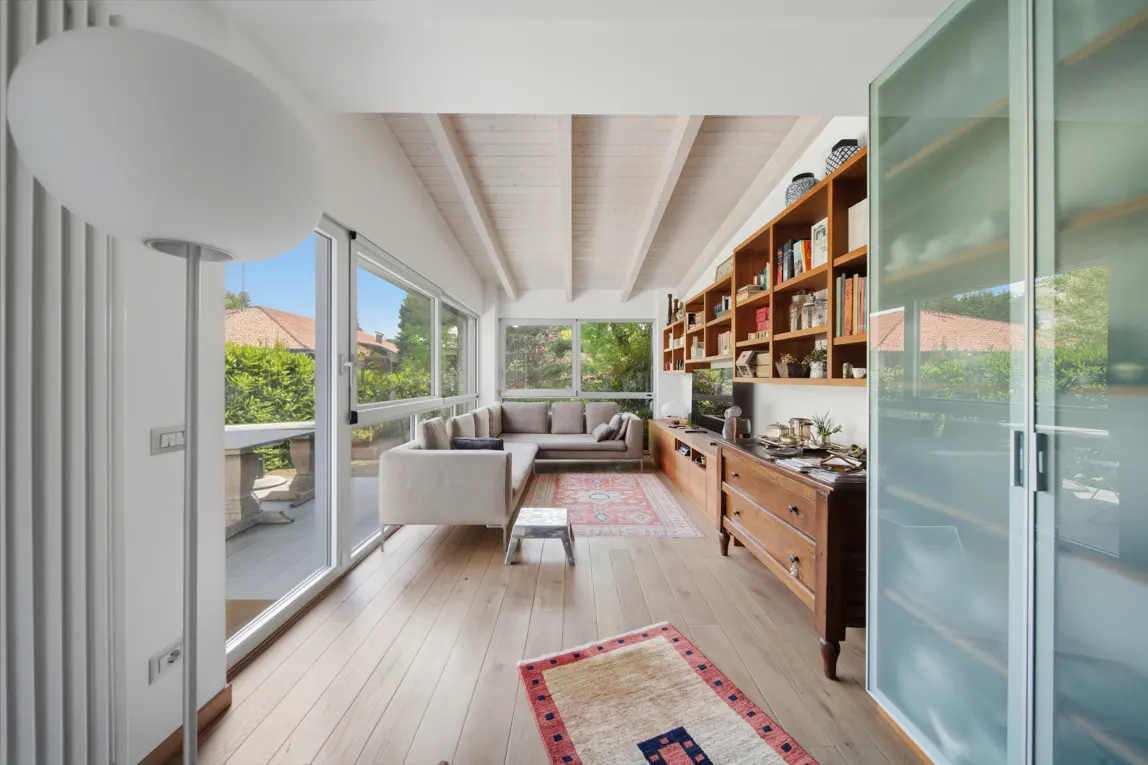 Bright living room with wood floors, white beamed ceiling, and large windows overlooking greenery. A gray sectional sofa sits near a small stool and patterned rugs. A wooden bookcase and frosted glass doors are visible.