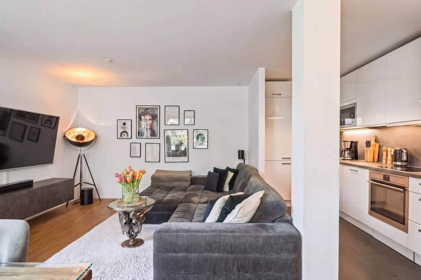 Open-concept living room with a gray sectional sofa, gallery wall, and a view into a modern white kitchen.