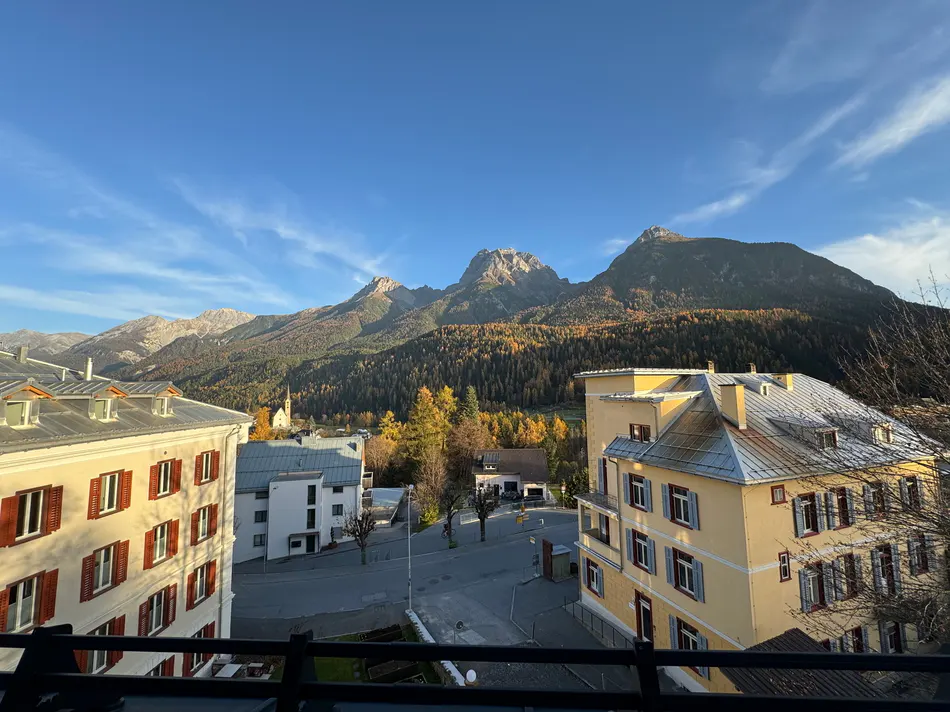 View from a balcony of yellow buildings, a street, and mountains with forests under a blue sky.