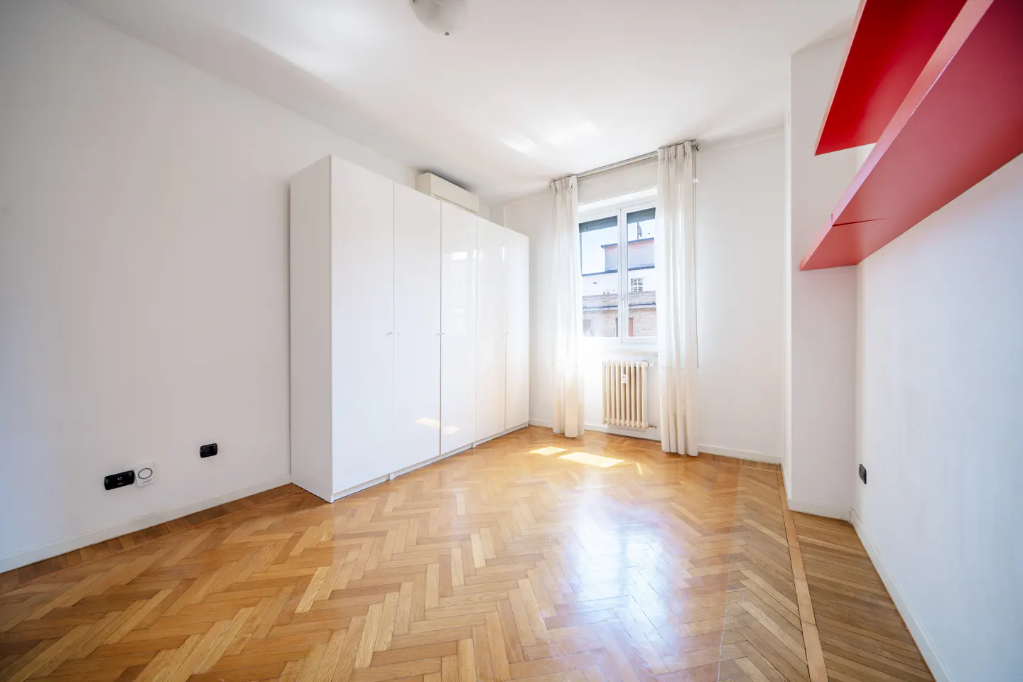 Bright, empty bedroom with herringbone wood floors, white walls, and a large white wardrobe. A window with sheer curtains lets in sunlight. Red shelves are on the right wall.
