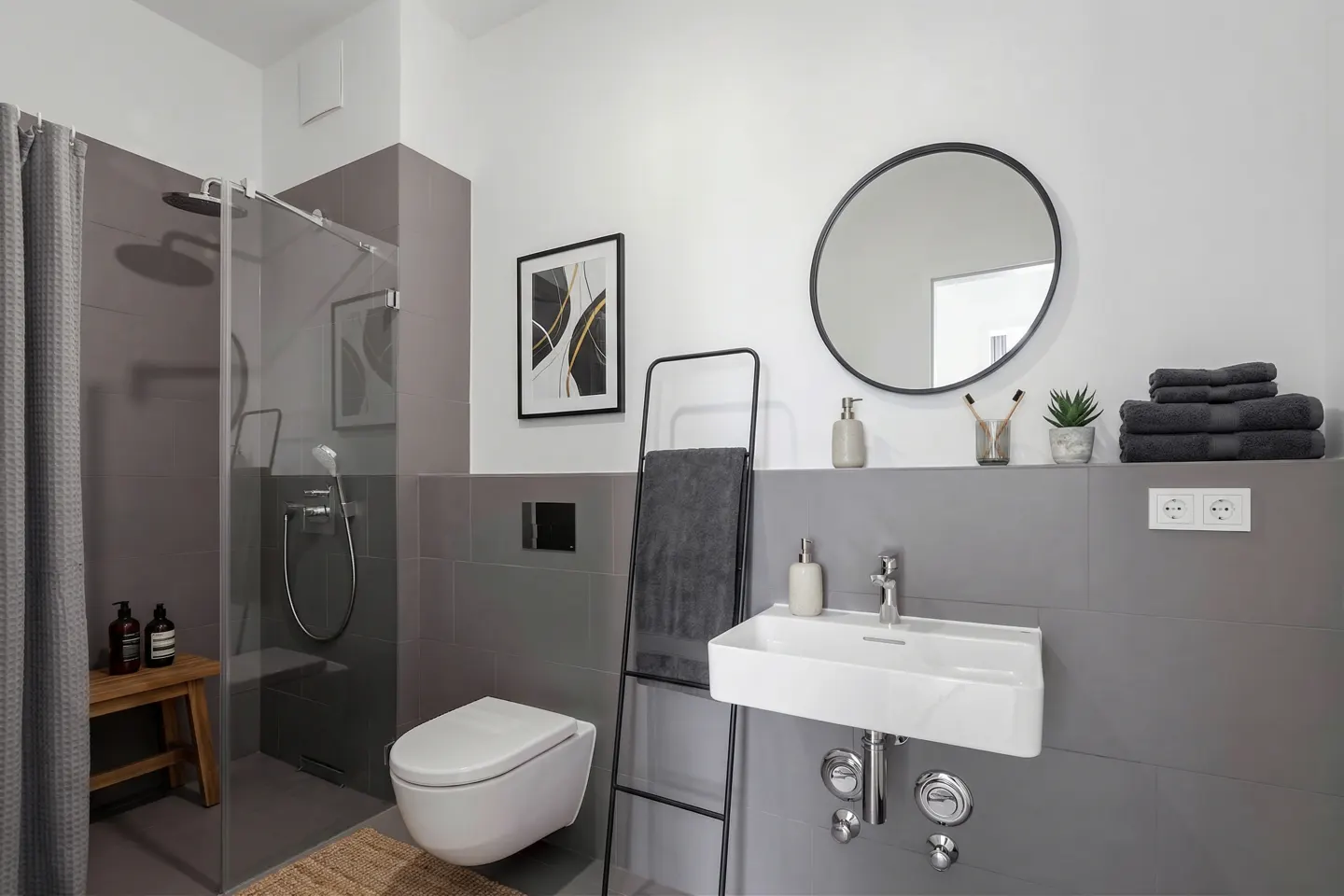Modern bathroom with gray tile, white toilet and sink, round mirror, and glass shower with gray curtain.