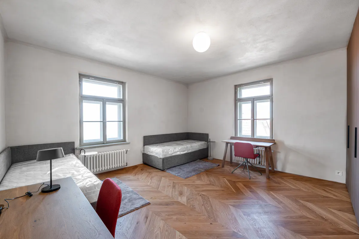 Bright bedroom with two beds, desks, and red chairs. Herringbone wood floors and white walls. Windows provide natural light.