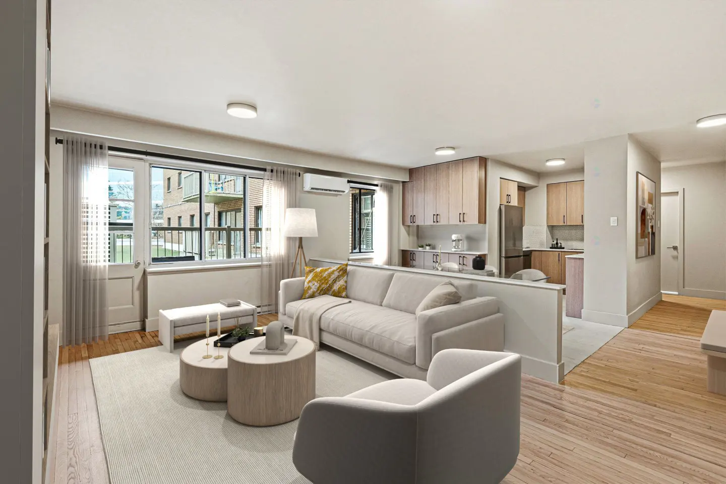 Bright, open-concept apartment with a beige sofa, round coffee tables, and a view of a brick building through a large window. The kitchen has light wood cabinets.