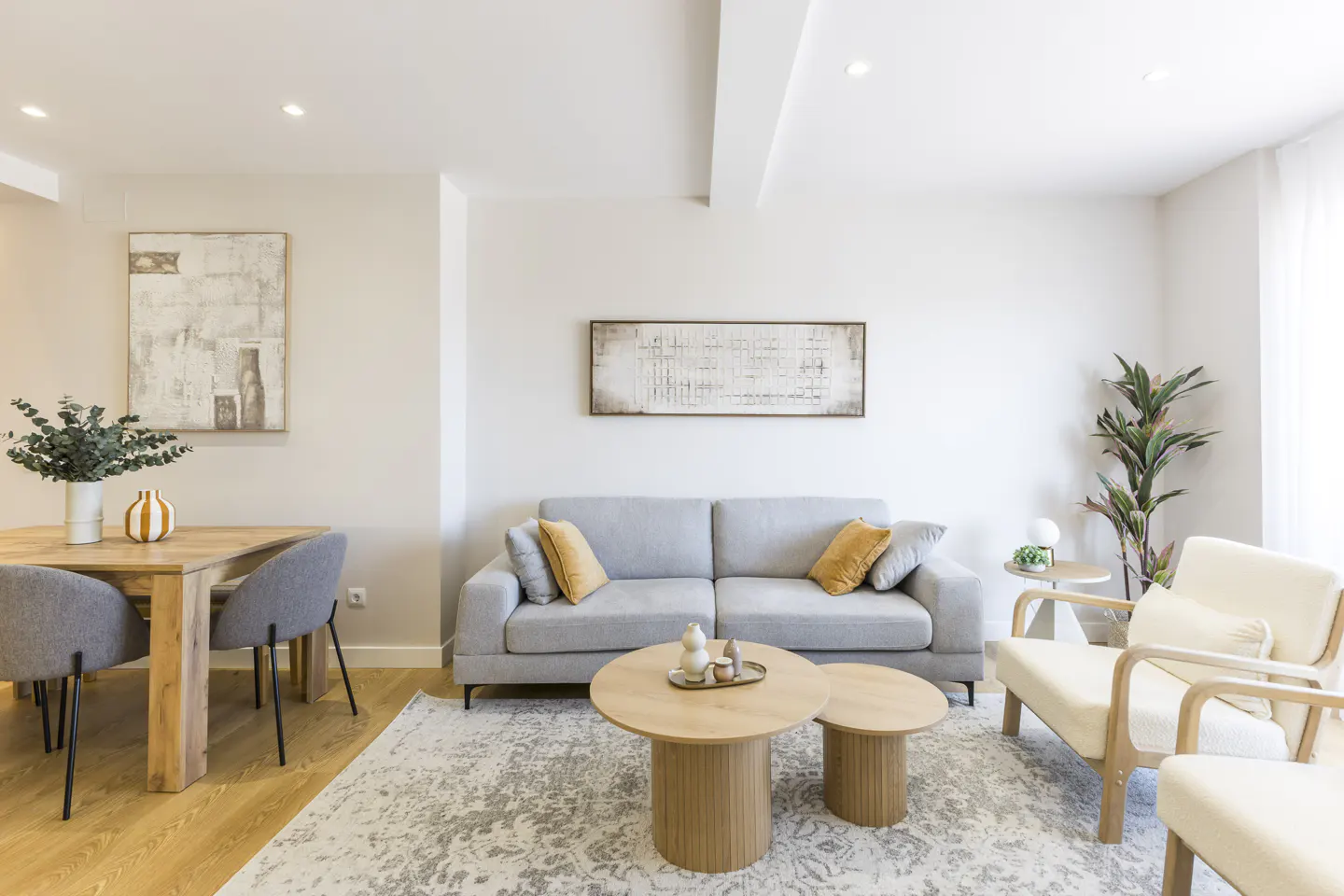 Bright living room with a gray sofa, two wooden coffee tables, and two white armchairs on a patterned rug. A wooden dining table with gray chairs is visible.