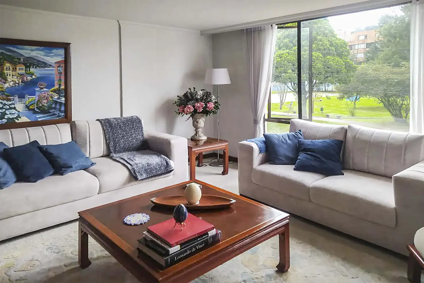 Bright living room with two beige sofas, blue pillows, a wooden coffee table, and a large window overlooking a park.
