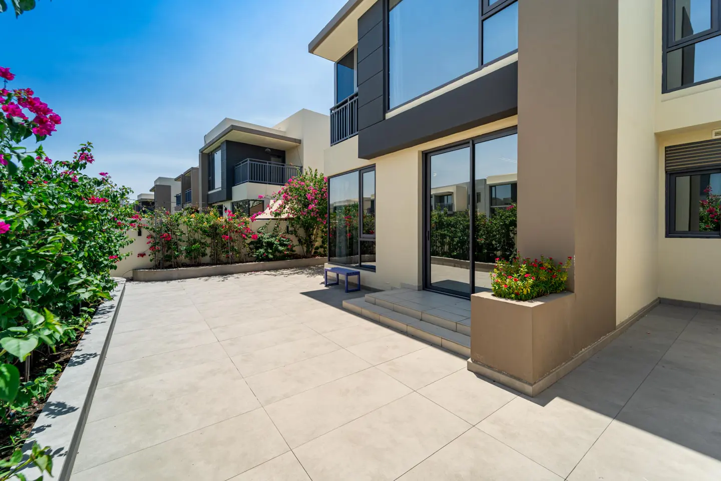 Exterior view of a modern beige house with large windows, a patio, and pink flowers under a blue sky.