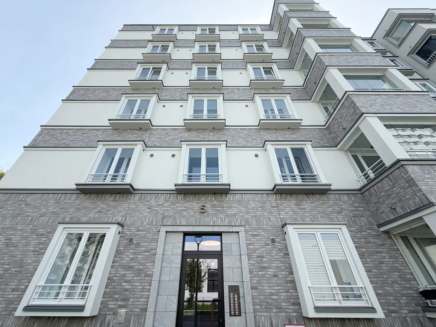 Modern apartment building with gray brick and white accents, viewed from below. Windows are white-framed and angled outward. The entrance door is black with glass panels.