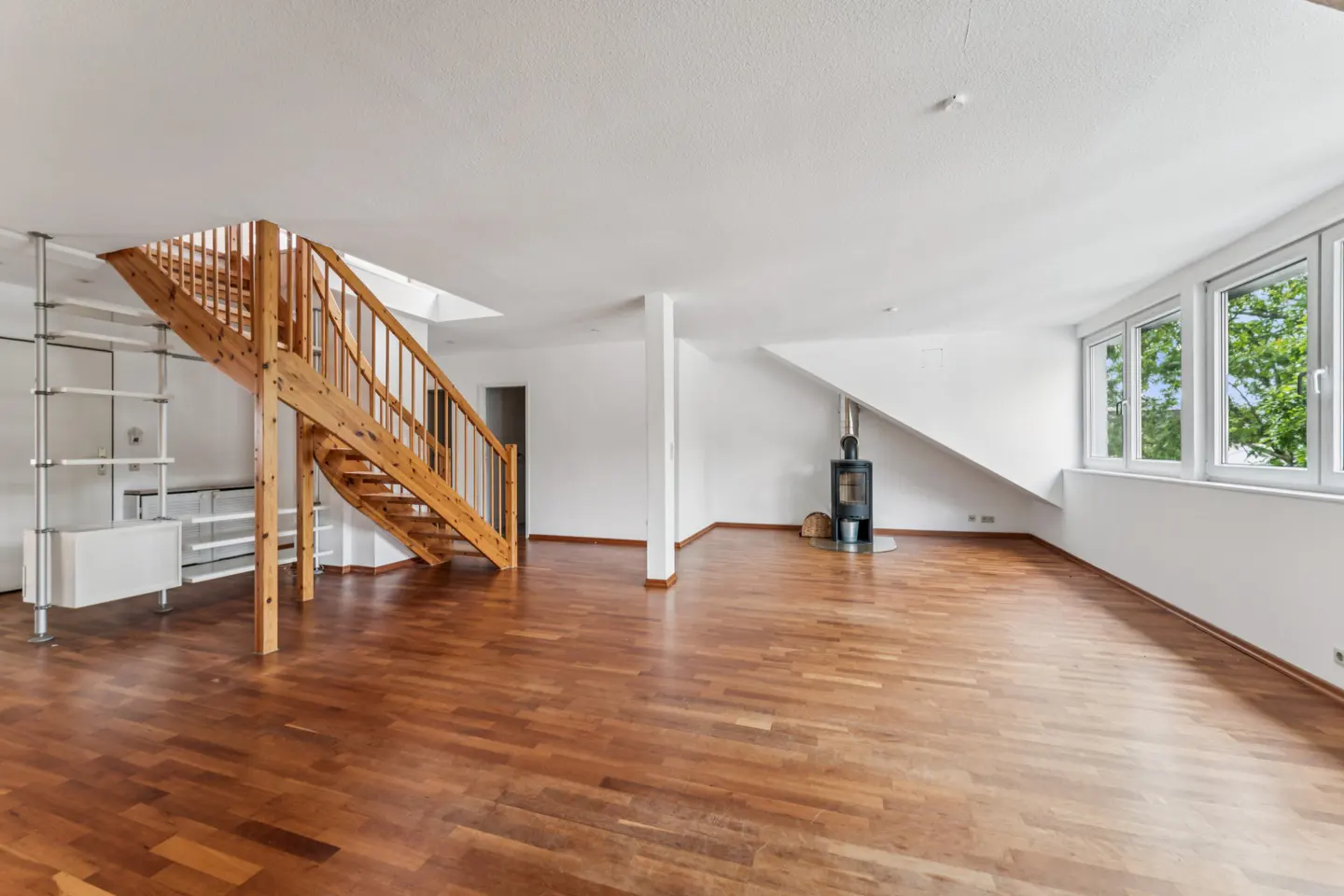 Spacious room with hardwood floors, a wooden staircase, a modern black fireplace, and large windows with white walls.