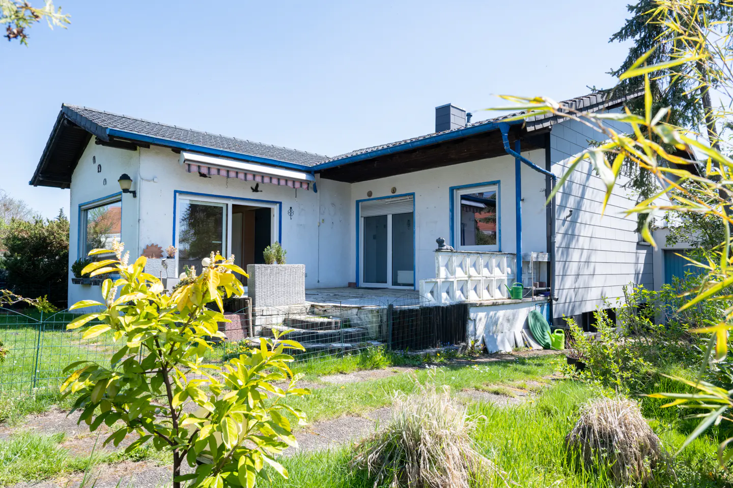 Exterior view of a one-story white house with blue trim, a gray roof, and a small patio with steps leading down to a grassy yard.
