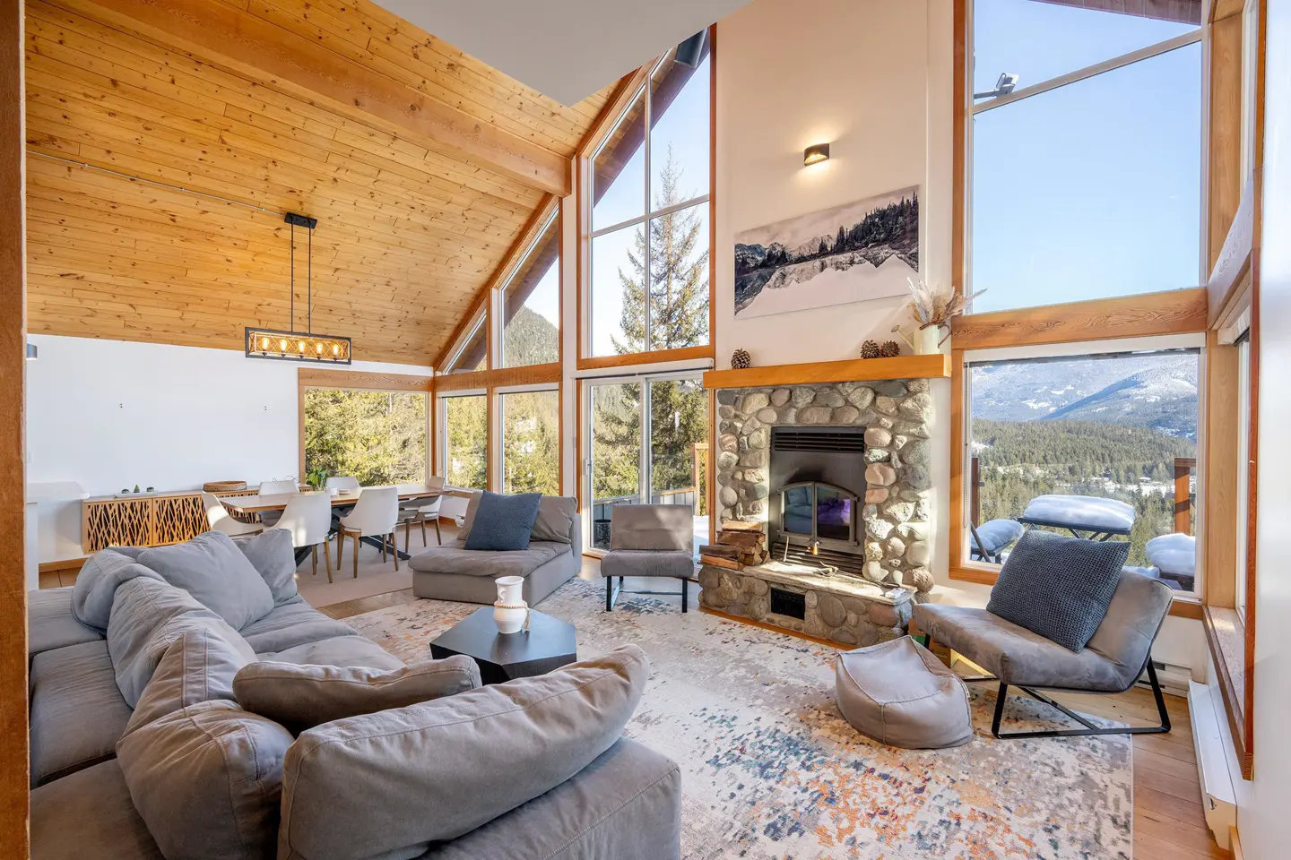 Bright living room with wood ceiling, stone fireplace, and large windows overlooking a snowy mountain landscape. Gray sectional sofa and dining table.