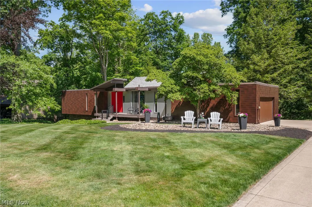 Mid-century modern home with brown siding, a bright red front door, and white patio chairs on a landscaped lawn.