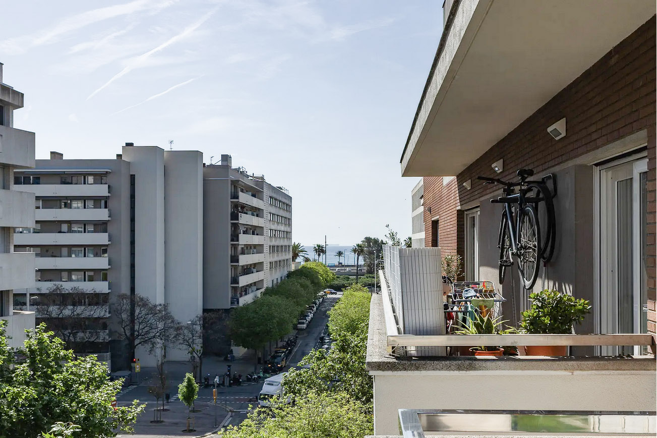 View from a balcony with a bicycle hanging on the wall, overlooking a city street with tall buildings and trees.
