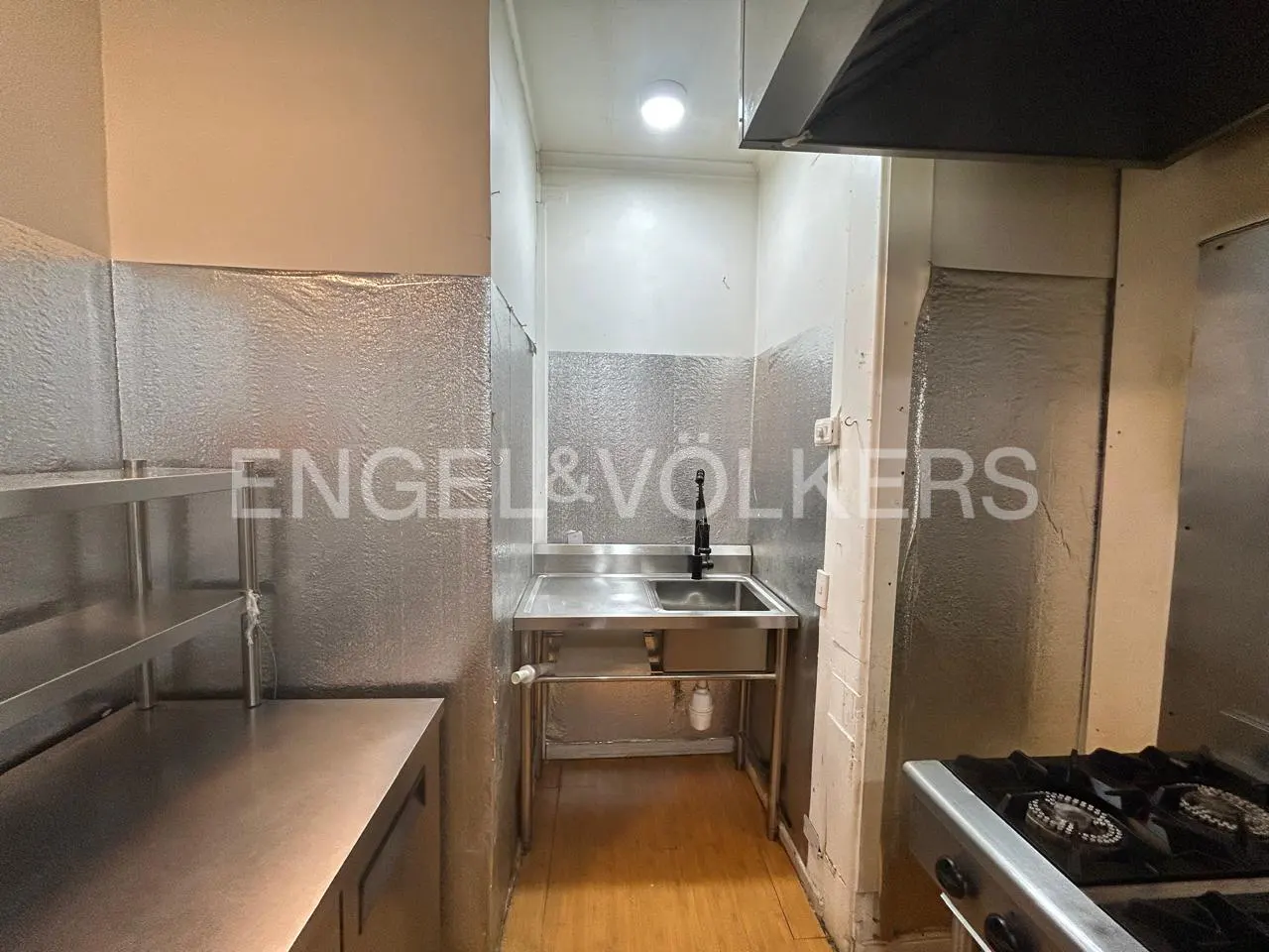 Commercial kitchen area with a stainless steel sink, metal wall panels, and a gas stovetop in the foreground.