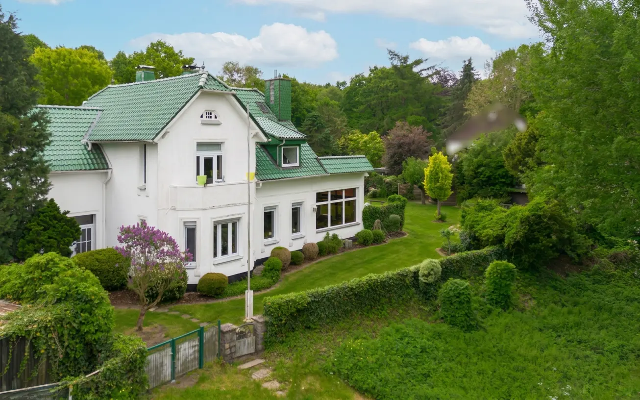 A white house with a green roof is surrounded by lush greenery and trees.