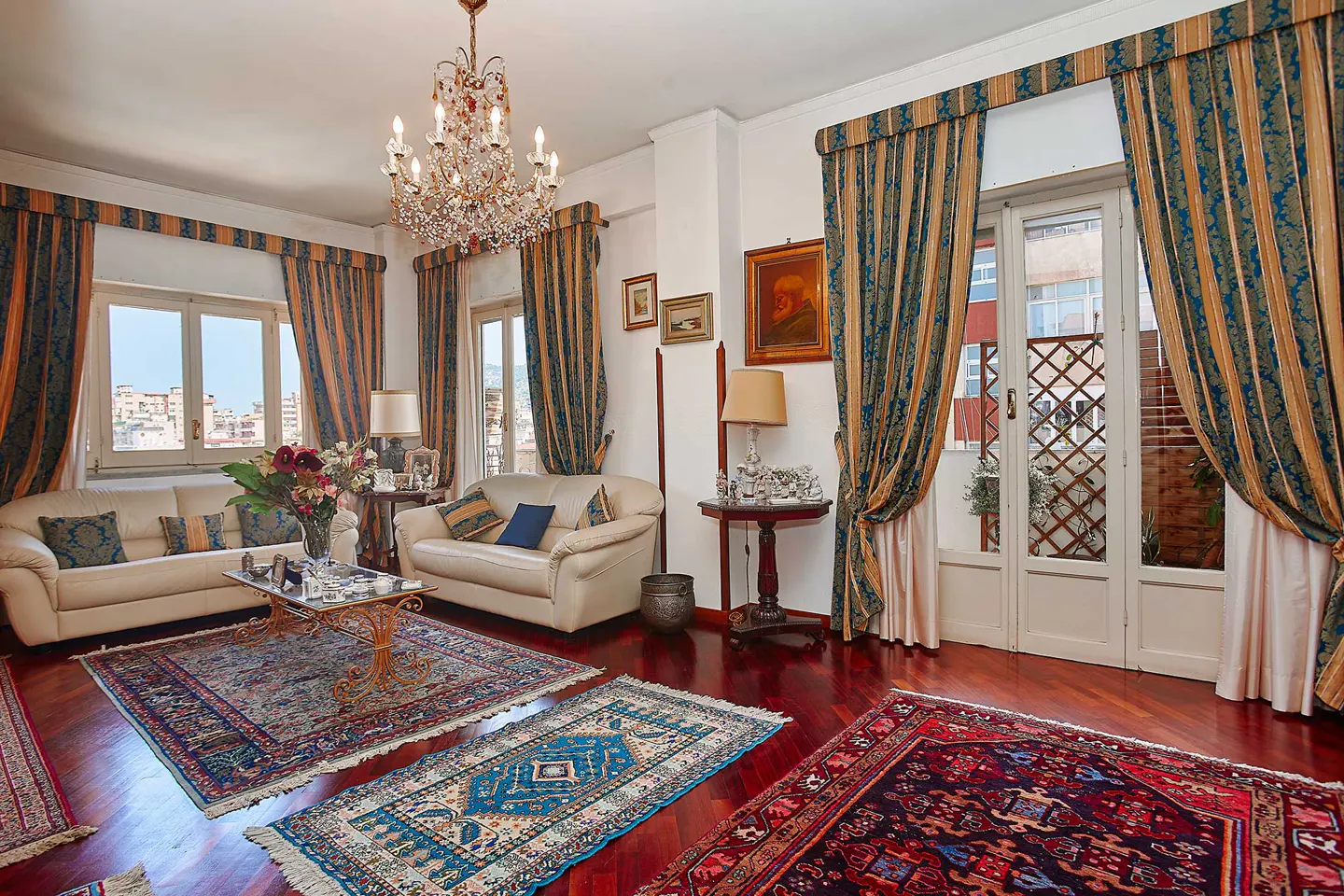 A living room with hardwood floors, two white sofas, and patterned rugs. A chandelier hangs from the ceiling. Blue and gold curtains frame the windows.