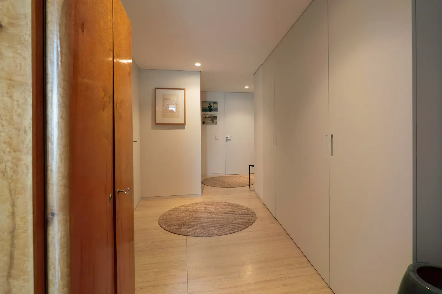 Hallway with light wood floors, white walls, and a wood cabinet. Round rugs are on the floor. Artwork hangs on the wall.