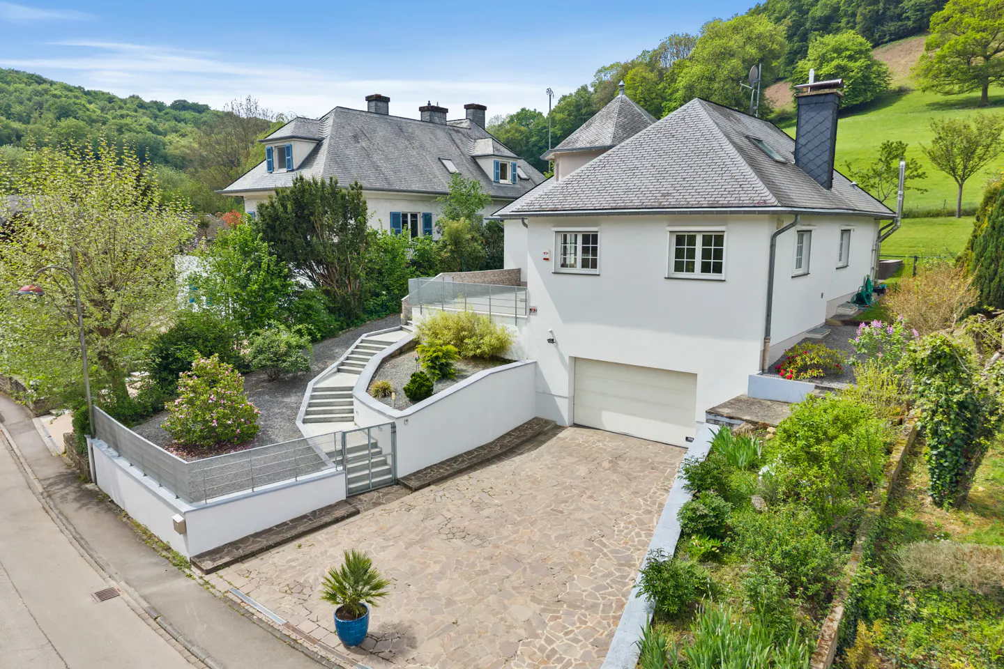 Aerial view of a white house with a gray roof, a garage, and a stone driveway surrounded by greenery.