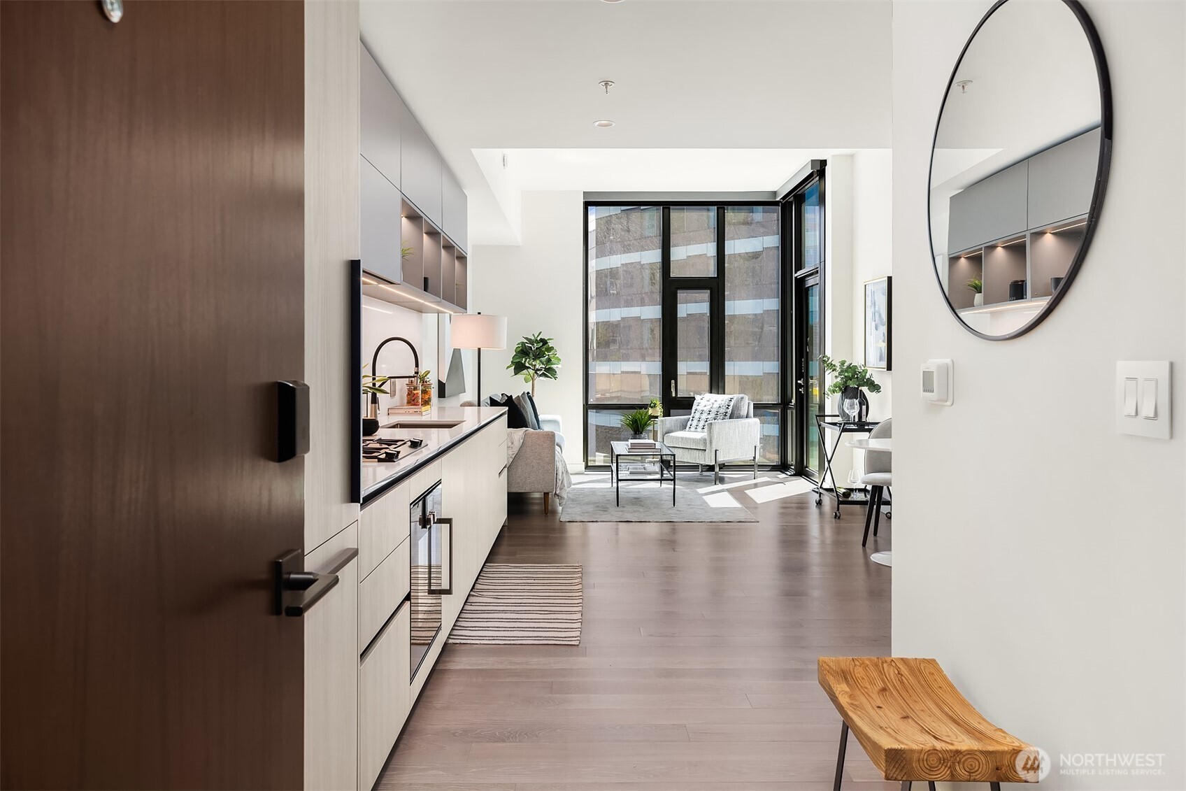 A modern apartment interior with a kitchen, living area, and hardwood floors. A round mirror hangs on the wall.